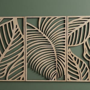 May include: Three-panel wooden wall art featuring a leaf design. The panels are made of light-colored wood and are cut out to create a negative space leaf pattern. The background is a muted green.