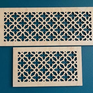 Può includere: Due coperture rettangolari in legno per bocchette di ventilazione con un motivo decorativo traforato. Il motivo presenta un design ripetuto di fiori a quattro petali e cerchi. Il legno è di colore chiaro e naturale, e lo sfondo è blu acqua.