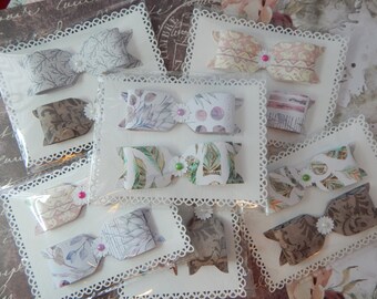 May include: A set of eight handmade paper bows, each with a different patterned design. The bows are displayed on white cards with lace trim and are packaged in clear plastic bags.