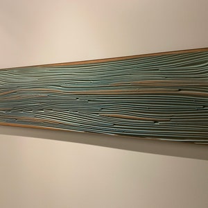 May include: A long, rectangular wall art piece with a wooden frame. The artwork is made up of many thin, layered strips of wood, painted in shades of blue, green, and brown. The strips are arranged in a wavy pattern, creating a sense of movement and depth.