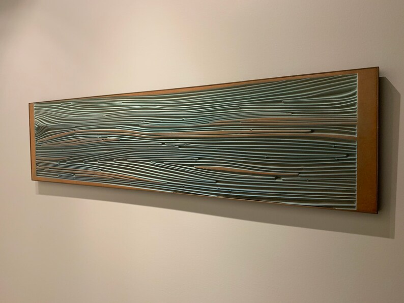May include: A long, rectangular wall art piece with a wooden frame. The artwork is made up of many thin, layered strips of wood, painted in shades of blue, green, and brown. The strips are arranged in a wavy pattern, creating a sense of movement and depth.