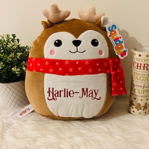 May include: A brown plush reindeer with a red and white snowflake patterned scarf. The reindeer has a white face with black eyes and a black nose. The reindeer is wearing a red scarf with the name "Harlie-May" embroidered on it.