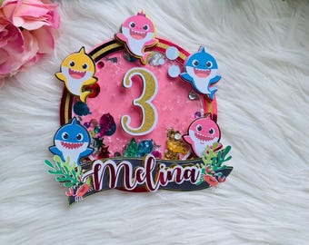 May include: A pink and gold glitter cake topper with the number 3 and the name "Melina". The topper features cartoon sharks in yellow, pink, and blue, with floral accents. The background is pink.