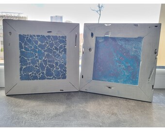 May include: Two square wall art pieces with distressed white wooden frames. The left piece features a blue and white mosaic tile pattern. The right piece displays a blue and purple fluid acrylic pour painting with visible marbled swirls and cells.