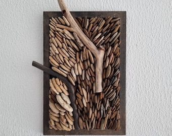 May include: A rectangular wall art piece featuring a mosaic of small, smooth, tan stones within a dark brown wooden frame. Natural driftwood branches are arranged across the stone mosaic, creating a textured, organic design. The artwork is mounted on a white wall.