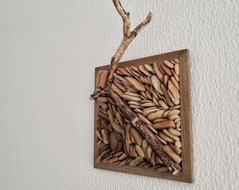 May include: A square, framed wall art piece featuring a mosaic of small, tan stones. A small, dried branch is affixed to the front, adding a natural element. The frame is made of wood, and the artwork is mounted on a white wall.