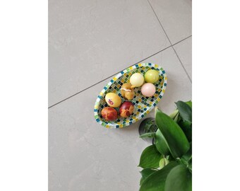 May include: An oval-shaped bowl featuring a mosaic pattern of small green, yellow, and blue tiles. Inside the bowl are several smooth, round objects including solid-colored spheres in yellow, cream, and pink, and two mottled brown and red patterned eggs.