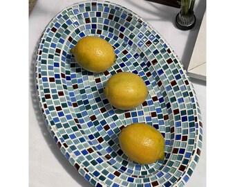 May include: An oval-shaped decorative tray featuring a mosaic pattern of small square tiles in various shades of blue, teal, and dark brown. Three bright yellow lemons are arranged in a line along the center of the tray's interior surface.