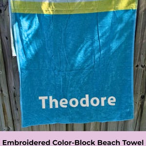 Könnte beinhalten: Ein türkisblaues Strandtuch mit einem weißen Streifen und einem gelben Streifen. Das Handtuch ist mit dem Namen "Theodore" in Weiß bestickt. Das Handtuch ist 91,44 cm x 182,88 cm groß.