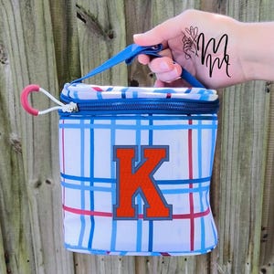 Puede incluir: Bolsa de almuerzo aislante a cuadros blancos y azules, con asa y cremallera azules. La bolsa presenta una gran letra "K" naranja y un patrón de cuadros rojos y azules. La bolsa está sujeta por una mano.