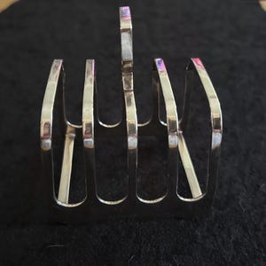 May include: A silver-toned metal toast rack featuring four vertical slots for holding slices of bread. The structure has a central handle loop at the top and a rectangular base, with a polished, reflective surface throughout the entire object.