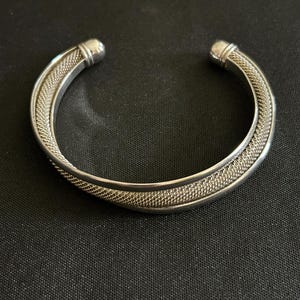 May include: A silver cuff bracelet with a mesh design. The bracelet has a rounded shape with a smooth, solid end cap on each side. The bracelet is displayed on a black surface.