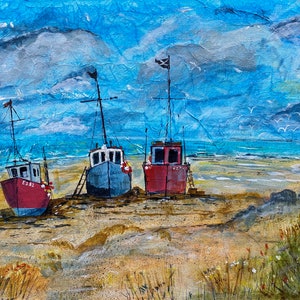 May include: Three small fishing boats are beached on a sandy shore with a blue sky and white clouds in the background. The boats are red, blue, and grey. The boats have flags on their masts.