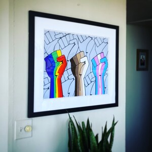May include: A framed print of three raised fists, each painted in a different color: rainbow, brown, and light blue with pink and white stripes. The fists are surrounded by a pattern of smaller fists.
