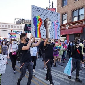 May include: A group of people marching in a street holding signs with the text "Black Trans Lives Matter" and a sign with three raised fists painted in rainbow, brown, and transgender pride colors.