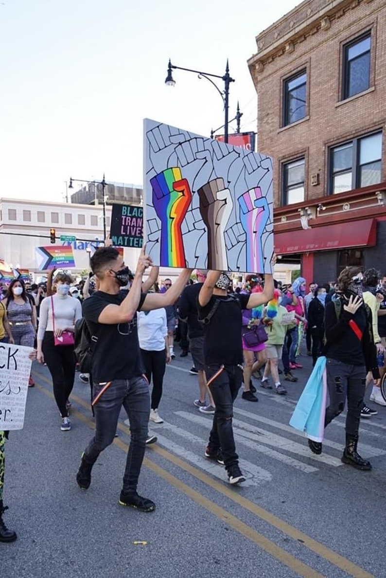 May include: A group of people marching in a street holding signs with the text "Black Trans Lives Matter" and a sign with three raised fists painted in rainbow, brown, and transgender pride colors.
