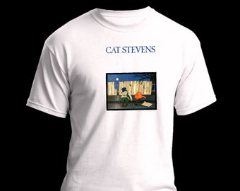 以下が含まれることがあります： 上部にネイビーブルーの「CAT STEVENS」の文字が入った白いTシャツ。その下に、フェンスの近くに座っている人物の額装されたイラストがあり、背景には満月があります。イラストの人物は帽子をかぶり、杖を持っています。