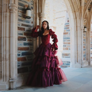 K&ouml;nnte beinhalten: Ein burgunderfarbenes Abendkleid mit einem geschichteten Rock und Puff&auml;rmeln. Das Kleid hat ein eng anliegendes Oberteil und einen mehrschichtigen Rock, der eine dramatische Silhouette erzeugt. Der Hintergrund umfasst Steinb&ouml;gen und Ziegelmauern.