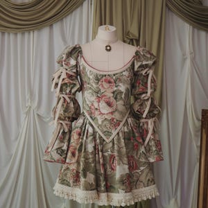 K&ouml;nnte beinhalten: Kleid mit floralem Muster mit rundem Ausschnitt und kurzem Rock. Das Kleid hat Puff&auml;rmel mit Schleifenbindungen und eine Spitzenborte. Der Stoff hat ein Muster aus rosa und roten Rosen auf gr&uuml;nem und cremefarbenem Hintergrund.