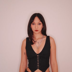 May include: A woman wearing a black corset top with a lace-up front and a black skirt with silver embellishments. She is wearing a silver necklace with a padlock pendant.
