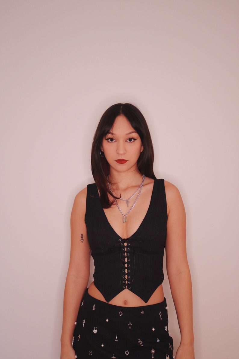 May include: A woman wearing a black corset top with a lace-up front and a black skirt with silver embellishments. She is wearing a silver necklace with a padlock pendant.