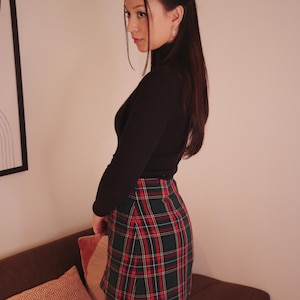 May include: A black turtleneck top paired with a red, green, and white plaid mini skirt. The skirt features a classic tartan pattern. The outfit is completed with black tights and a hair clip. The overall style is preppy and chic.