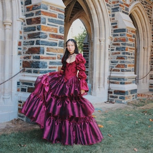 K&ouml;nnte beinhalten: Ein burgunderfarbenes Ballkleid mit einem figurbetonten Oberteil und einem geschichteten, ger&uuml;schten Rock. Das Kleid hat lange, gepuffte &Auml;rmel und einen hohen Ausschnitt. Das Kleid wird im Freien in der N&auml;he eines Steingeb&auml;udes mit gew&ouml;lbten T&uuml;ren getragen.