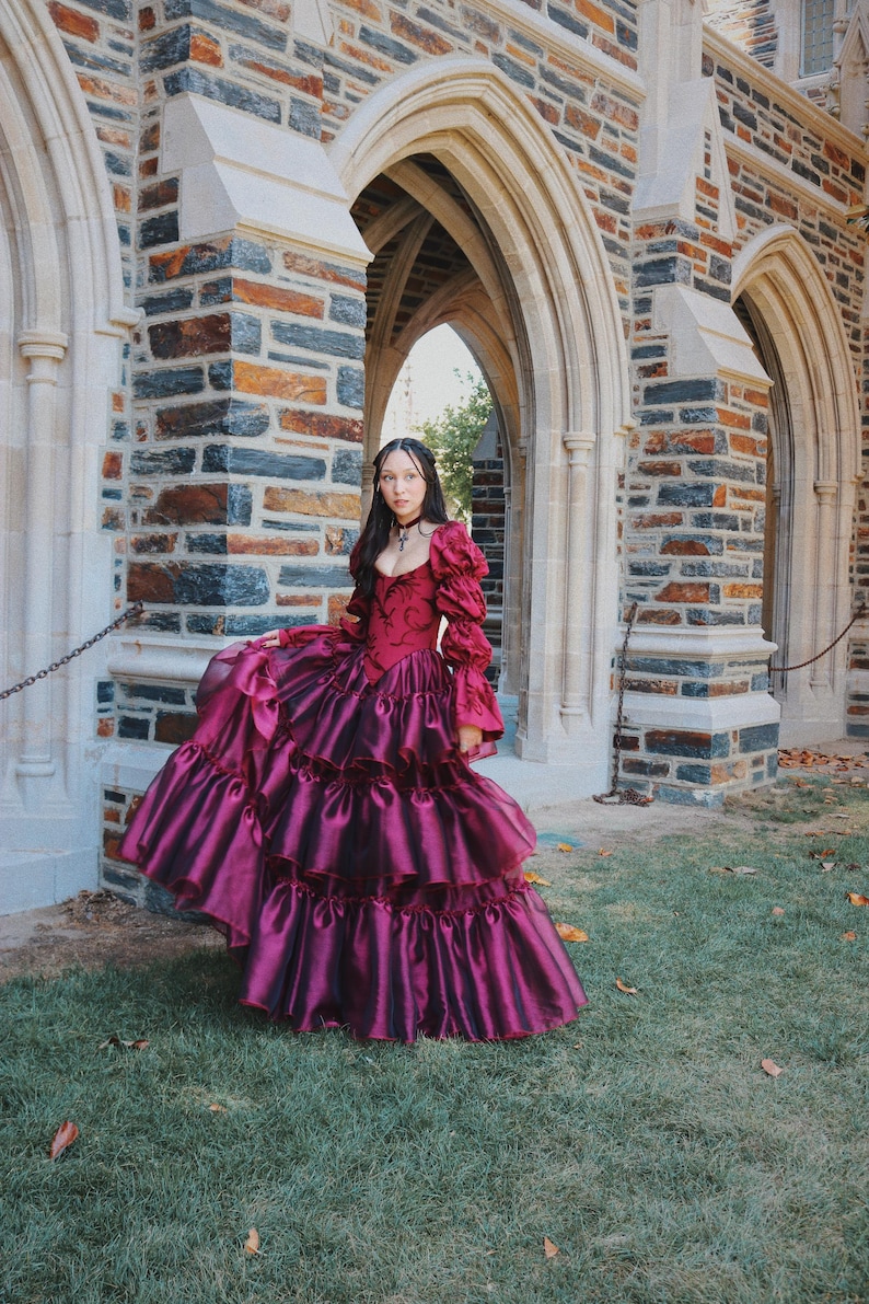 K&ouml;nnte beinhalten: Ein burgunderfarbenes Ballkleid mit einem figurbetonten Oberteil und einem geschichteten, ger&uuml;schten Rock. Das Kleid hat lange, gepuffte &Auml;rmel und einen hohen Ausschnitt. Das Kleid wird im Freien in der N&auml;he eines Steingeb&auml;udes mit gew&ouml;lbten T&uuml;ren getragen.