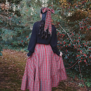 May include: A long, red and gray plaid skirt with a tiered ruffle hem. The person is wearing a black sweater and a matching plaid bow in their hair. The scene is set outdoors with trees and greenery in the background.