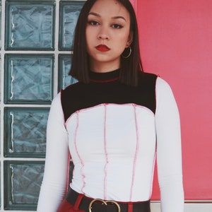 May include: A woman wearing a white and black long-sleeved shirt with pink stitching, red pants, and a black belt with a gold buckle. She is standing in front of a red door and a glass block wall.
