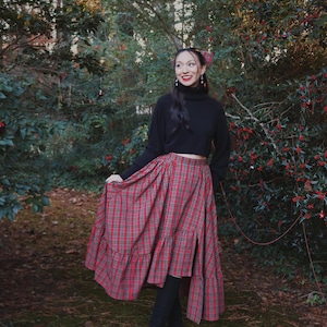 May include: A red and green plaid skirt with a tiered ruffle design. The skirt is paired with a black turtleneck sweater and black boots. The outfit is set in a natural outdoor setting with trees and greenery.