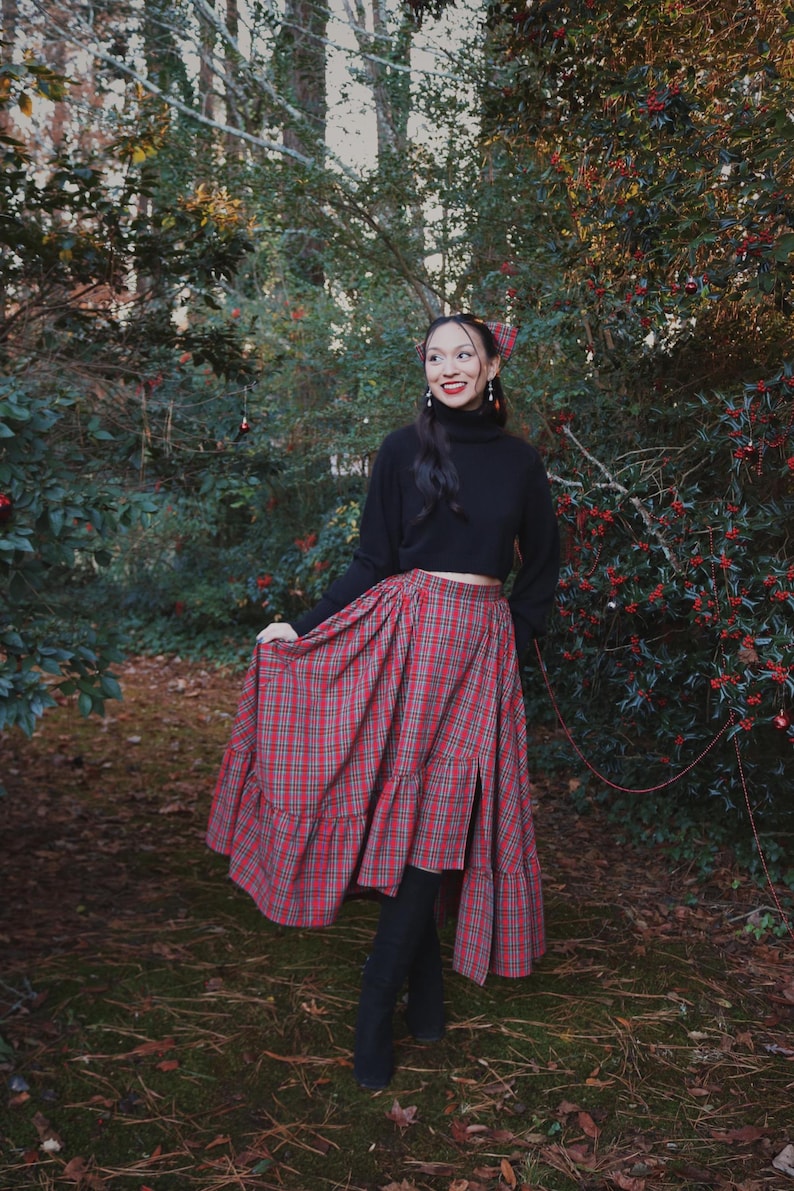 May include: A red and green plaid skirt with a tiered ruffle design. The skirt is paired with a black turtleneck sweater and black boots. The outfit is set in a natural outdoor setting with trees and greenery.