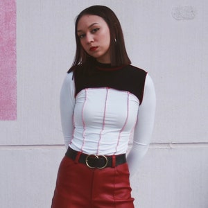 May include: A woman wearing a white and black long-sleeved shirt with red stitching and a black belt with a gold double-ring buckle. She is wearing red leather pants.