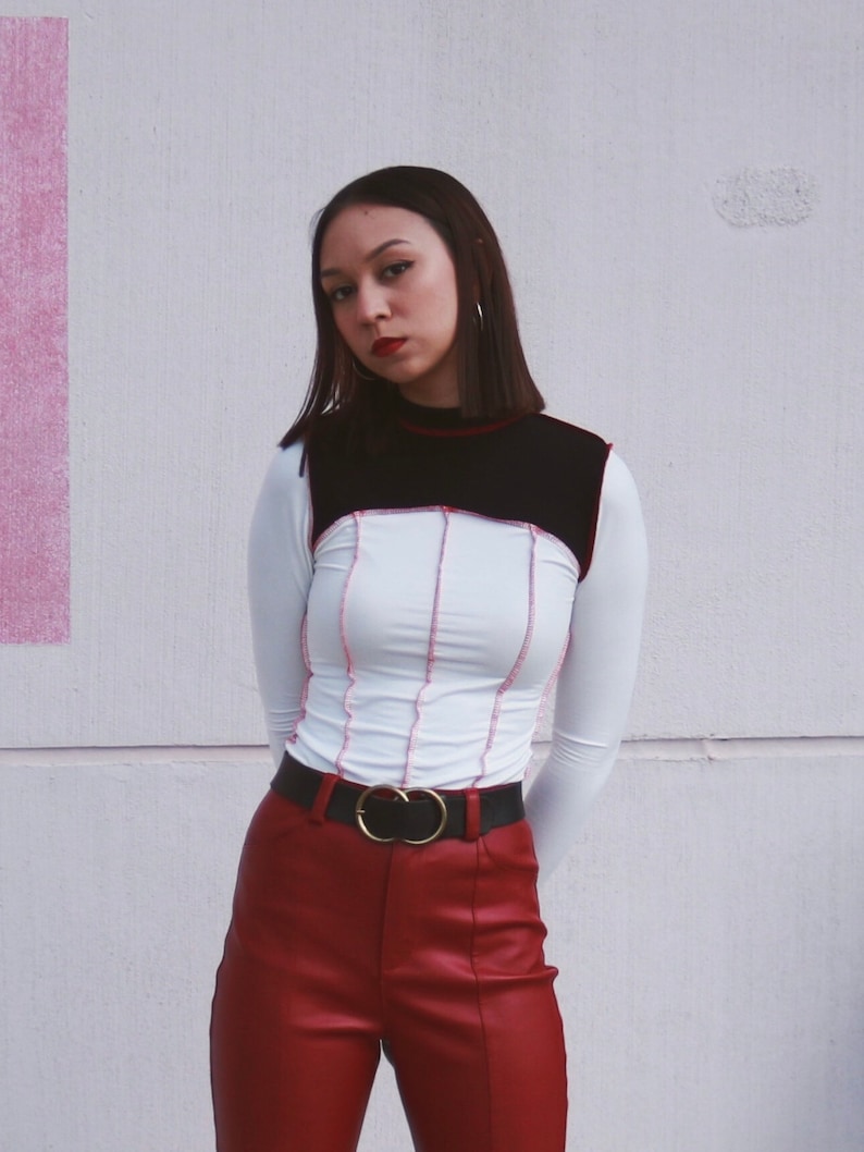 May include: A woman wearing a white and black long-sleeved shirt with red stitching and a black belt with a gold double-ring buckle. She is wearing red leather pants.