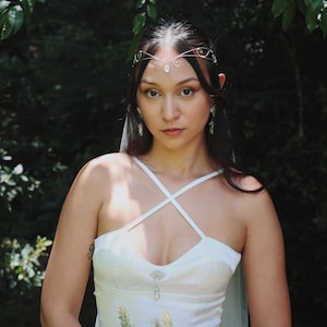 May include: A woman wearing a white wedding dress with a crisscross bodice, holding a bouquet of pink and white flowers. She is adorned with a delicate silver headpiece and earrings. The background is a lush green forest.