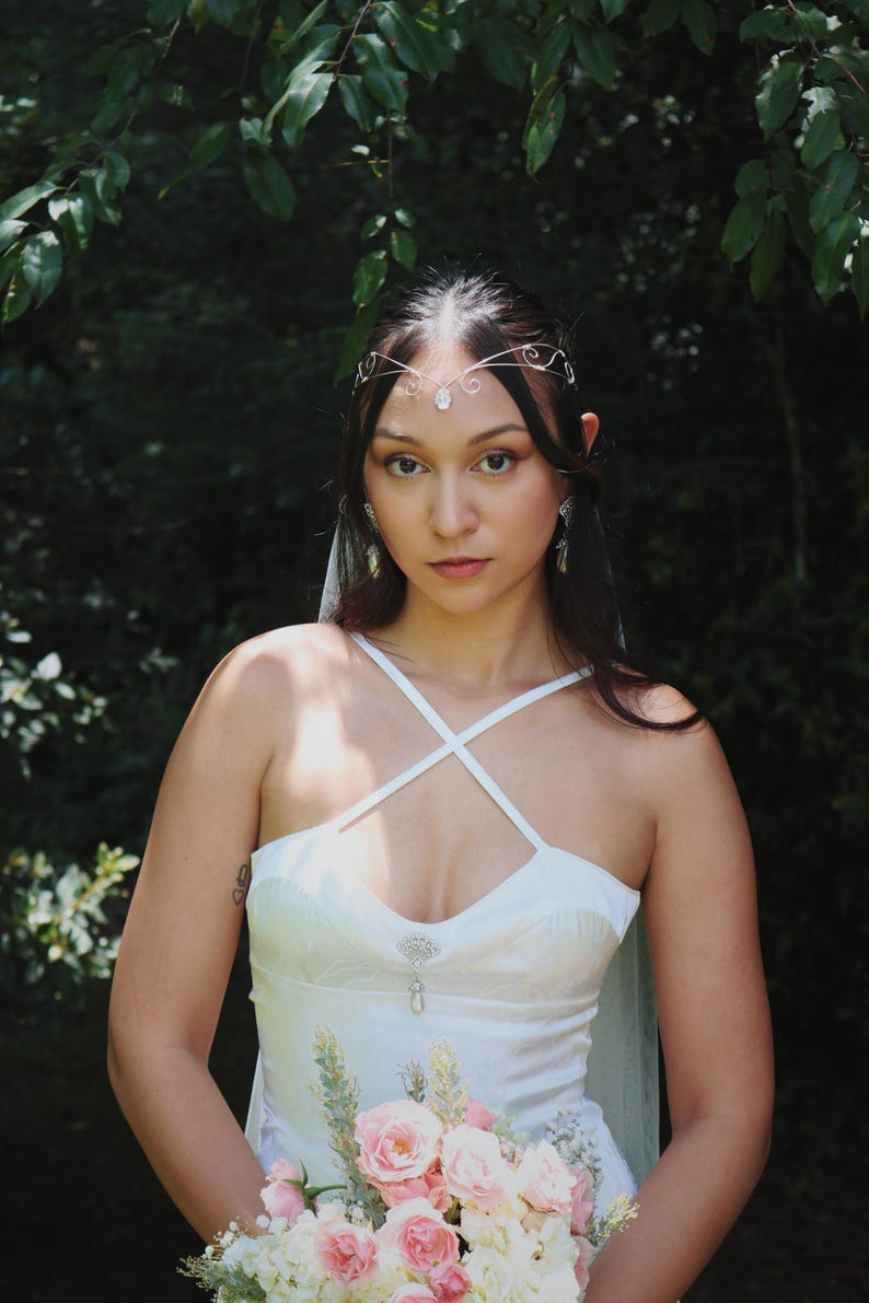 May include: A woman wearing a white wedding dress with a crisscross bodice, holding a bouquet of pink and white flowers. She is adorned with a delicate silver headpiece and earrings. The background is a lush green forest.