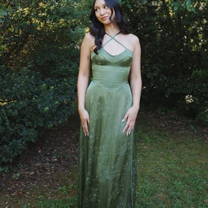 May include: A long, olive green dress with a floral pattern. The dress features a halter neckline with criss-cross straps and a fitted bodice that flows into a floor-length skirt. The dress is made of a silky material and is worn outdoors.