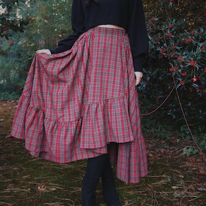 May include: A long, red plaid skirt with a tiered ruffle hem. The skirt is paired with a black turtleneck sweater and black boots. The skirt's plaid pattern includes red, green, and gray lines. The outfit is worn outdoors in a natural setting.