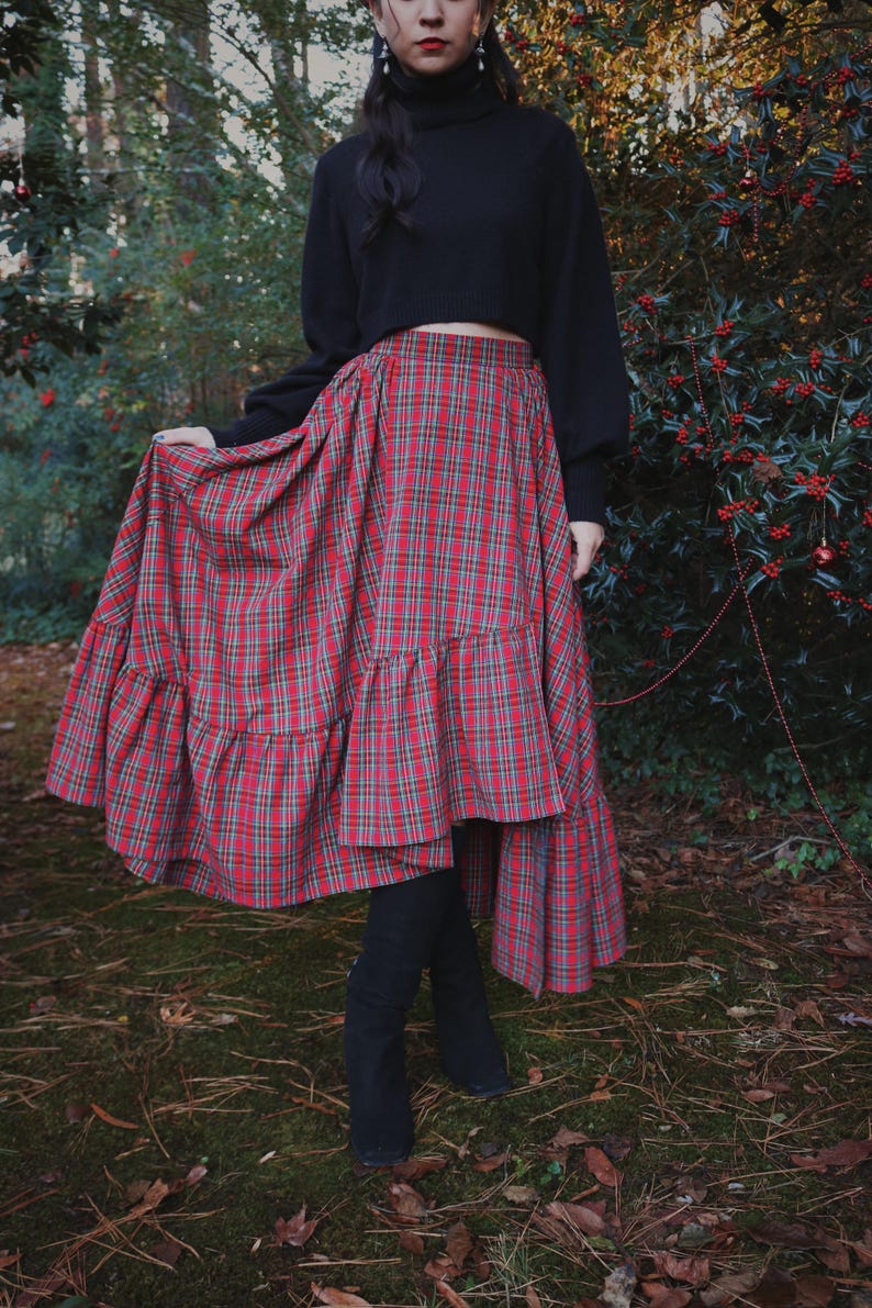 May include: A long, red plaid skirt with a tiered ruffle hem. The skirt is paired with a black turtleneck sweater and black boots. The skirt's plaid pattern includes red, green, and gray lines. The outfit is worn outdoors in a natural setting.