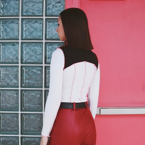 May include: A white long-sleeved shirt with black and red stitching detail. The shirt has a black panel on the back. The person is wearing red pants and a black belt.