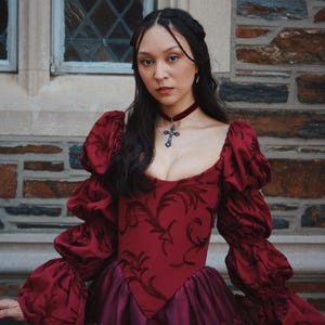 K&ouml;nnte beinhalten: Burgunderfarbenes Kleid mit herzf&ouml;rmigem Ausschnitt und Puff&auml;rmeln. Das Oberteil hat ein dunkles Blumenmuster. Das Kleid hat einen mehrschichtigen Rock in einem dunkleren Burgunderfarbton. Ein Samtchoker mit Kreuzanh&auml;nger vervollst&auml;ndigt den Look.