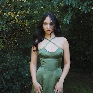 May include: A green satin dress with a crisscross halter neckline. The dress has a fitted bodice and a flowing skirt. The model is standing in front of a lush green background of trees and foliage. The dress has a subtle floral pattern.
