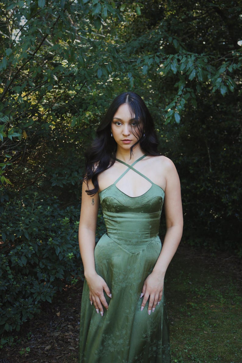May include: A green satin dress with a crisscross halter neckline. The dress has a fitted bodice and a flowing skirt. The model is standing in front of a lush green background of trees and foliage. The dress has a subtle floral pattern.