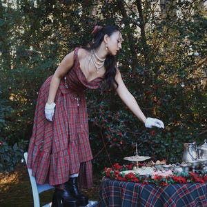 May include: A woman in a red plaid dress and black platform boots stands on a light blue chair, reaching towards a table set for tea. The table is covered with a matching plaid tablecloth and adorned with silver serving pieces and red floral decorations.
