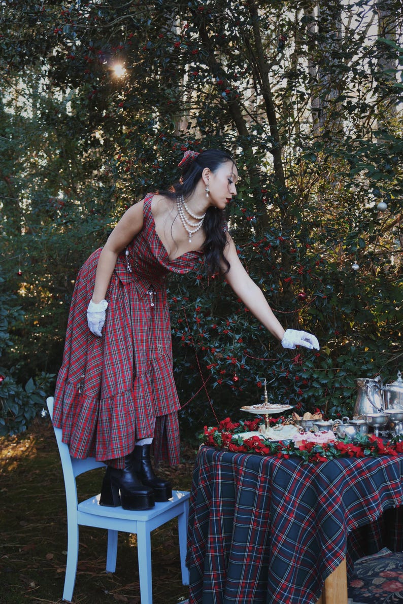 May include: A woman in a red plaid dress and black platform boots stands on a light blue chair, reaching towards a table set for tea. The table is covered with a matching plaid tablecloth and adorned with silver serving pieces and red floral decorations.