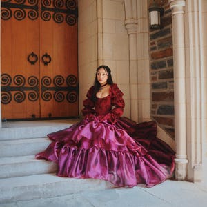 K&ouml;nnte beinhalten: Eine Person in einem bordeauxroten und magentafarbenen Ballkleid sitzt auf Steinstufen. Das Kleid hat einen ger&uuml;schten Rock und lange &Auml;rmel. Der Hintergrund umfasst eine Holzt&uuml;r mit Eisenarbeiten und einen Steinbogen.
