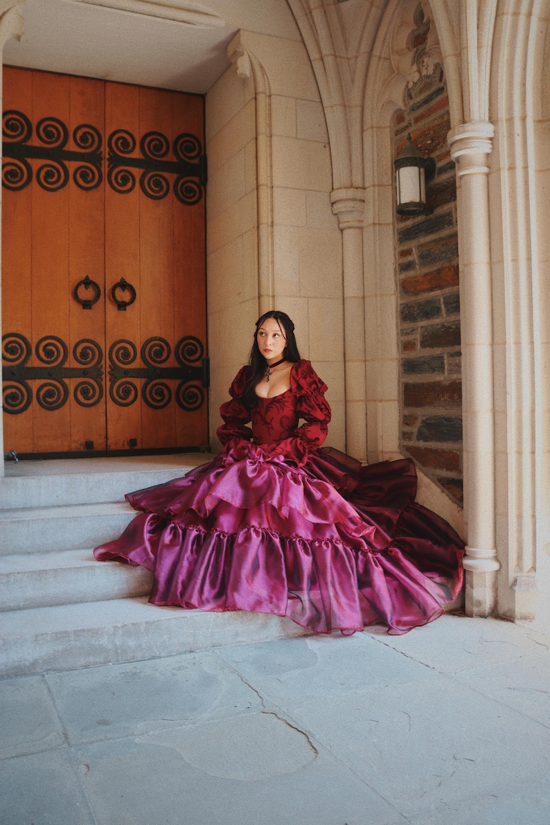 K&ouml;nnte beinhalten: Eine Person in einem bordeauxroten und magentafarbenen Ballkleid sitzt auf Steinstufen. Das Kleid hat einen ger&uuml;schten Rock und lange &Auml;rmel. Der Hintergrund umfasst eine Holzt&uuml;r mit Eisenarbeiten und einen Steinbogen.