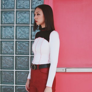 May include: A woman wearing a white long-sleeved shirt with black and pink stitching, a black belt, and burgundy pants. She is standing in front of a wall with a glass block window.