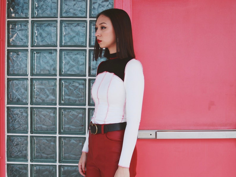 May include: A woman wearing a white long-sleeved shirt with black and pink stitching, a black belt, and burgundy pants. She is standing in front of a wall with a glass block window.