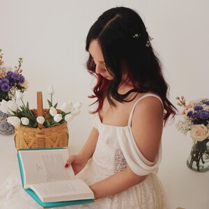 K&ouml;nnte beinhalten: Eine Frau in einem wei&szlig;en Spitzenkleid mit einem Blumenhaarband liest ein Buch mit t&uuml;rkisfarbenem Einband. Sie sitzt auf einem Stapel B&uuml;cher mit Blumen in Vasen auf beiden Seiten von ihr.